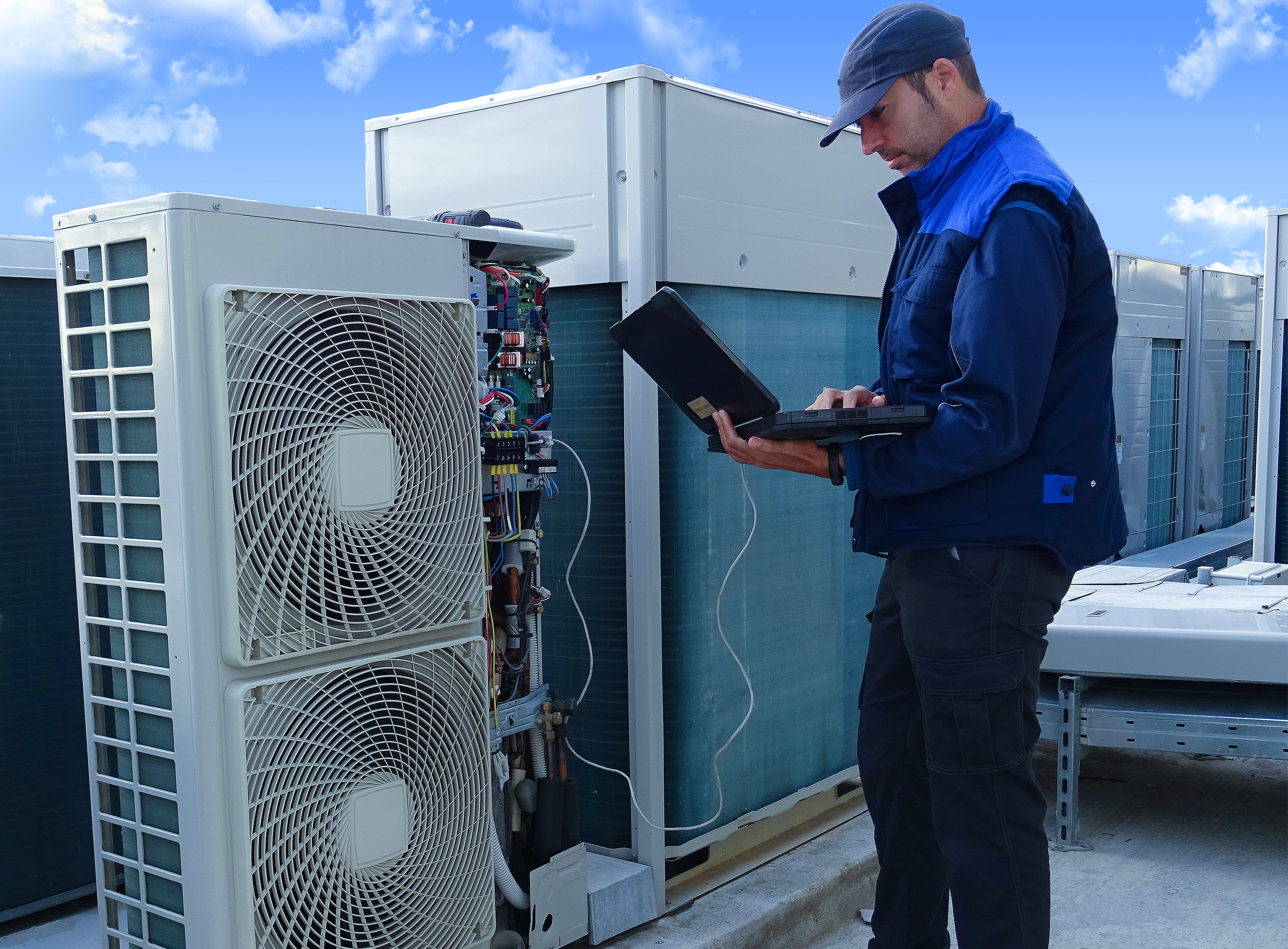 air conditioning technician making a diagnosis of an industrial air conditioning unit with a laptop next to other VRV condenser units on a rooftop in a sunny day