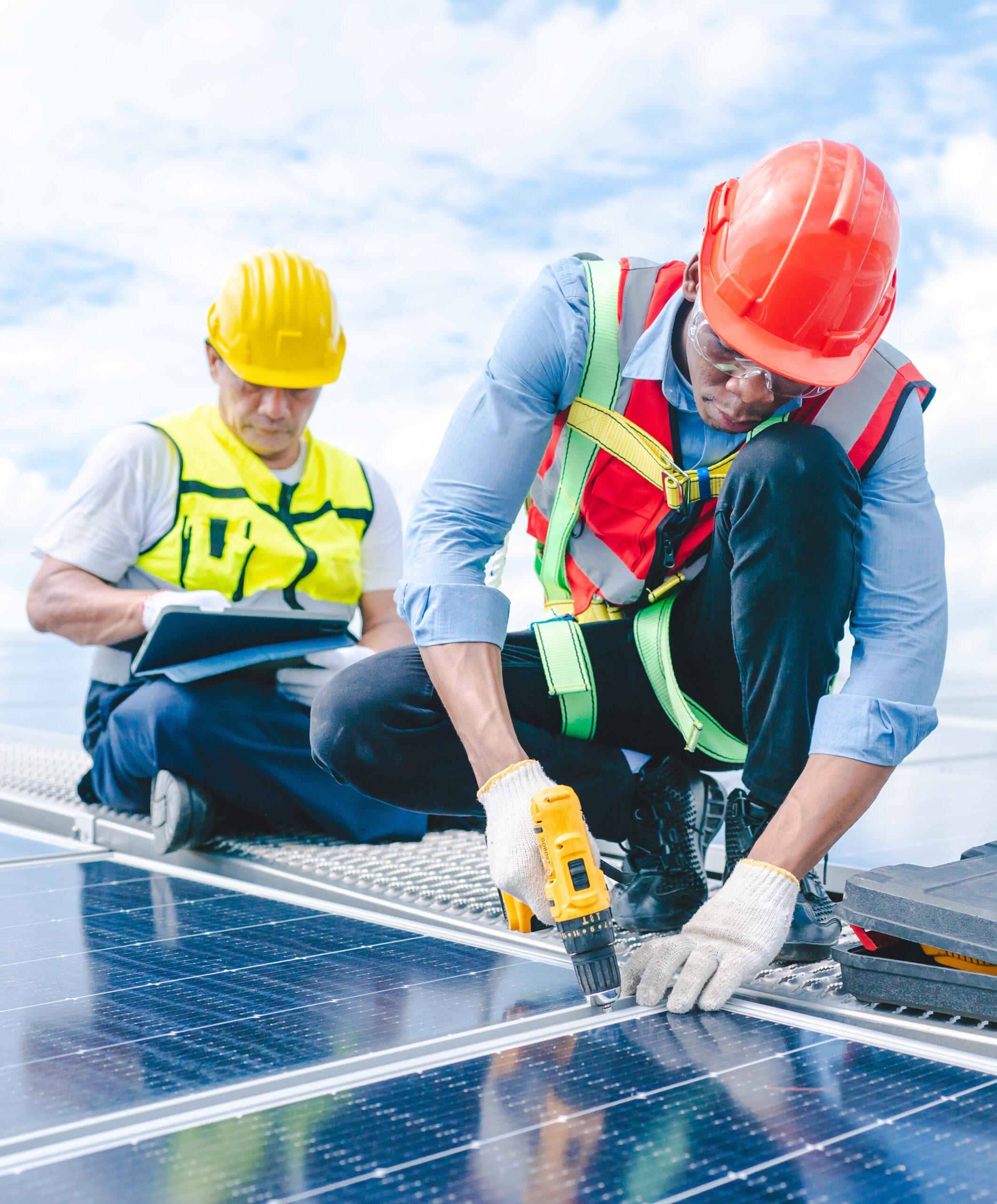 Two men in work hats and safety vests on a roof, one is using a drill on a solar panel, while the other is working on a tablet.